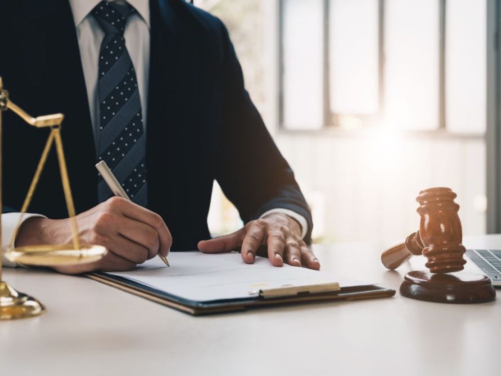 Lawyer signing documents at desk with gavel, scales, and laptop, symbolizing legal work and justice.