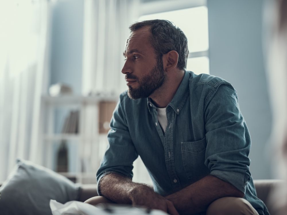 Man in denim shirt sitting thoughtfully in a well-lit room.