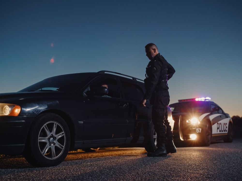 Police officer conducts traffic stop beside a car at dusk, with patrol lights illuminating the scene.