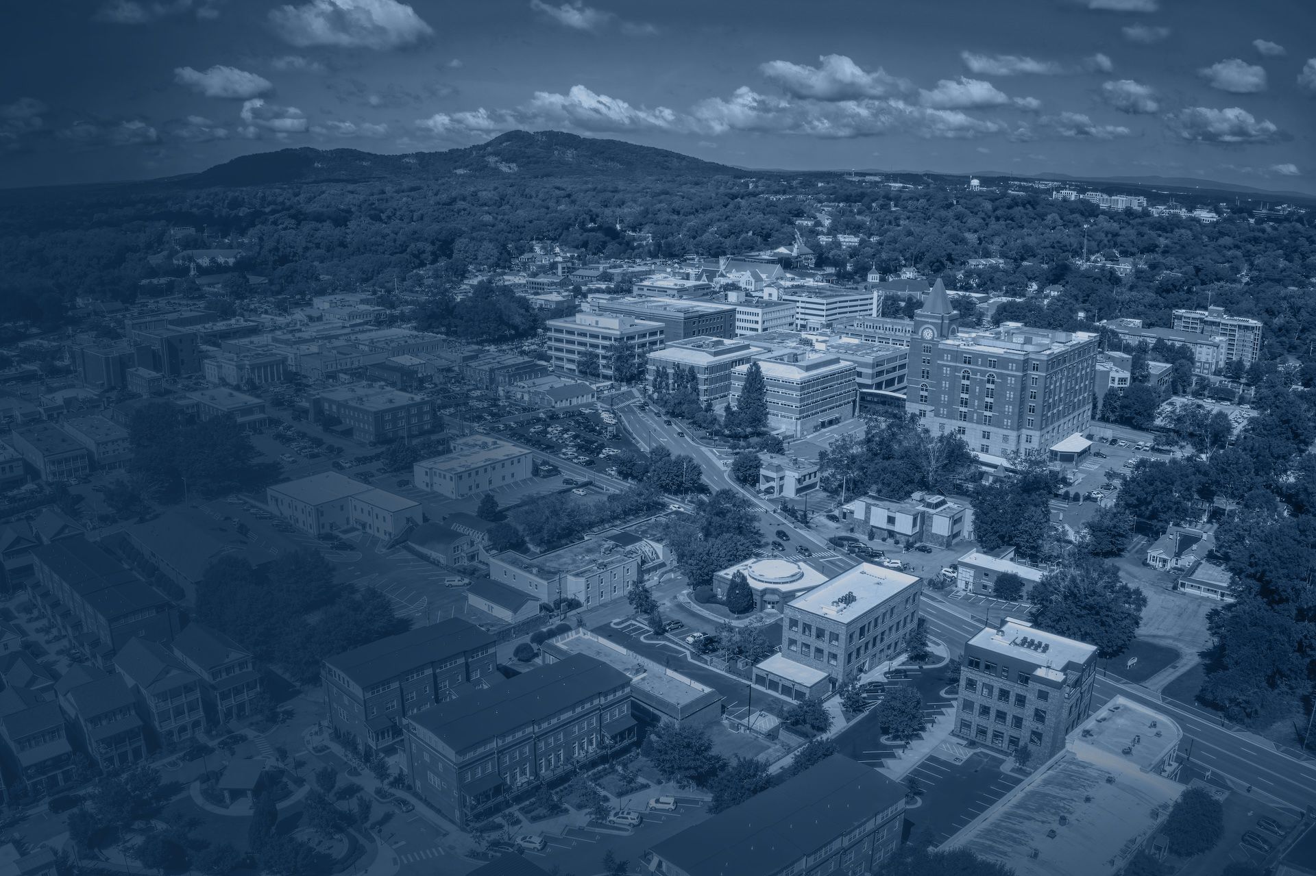 Aerial view of a cityscape with buildings, roads, and mountains in the background under a cloudy sky.