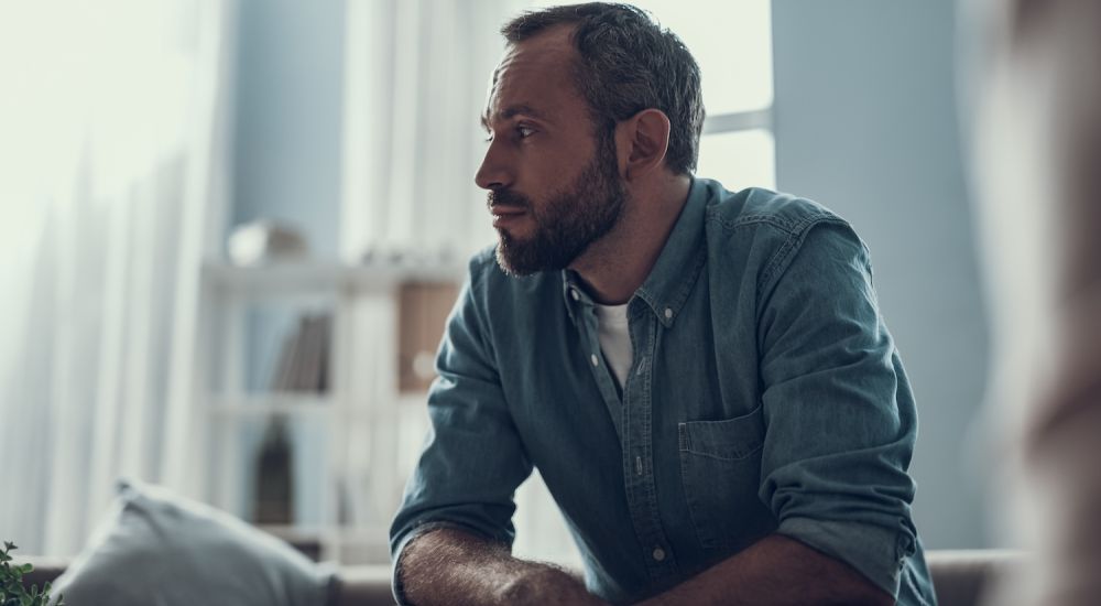 Man in denim shirt sitting thoughtfully in a well-lit room.