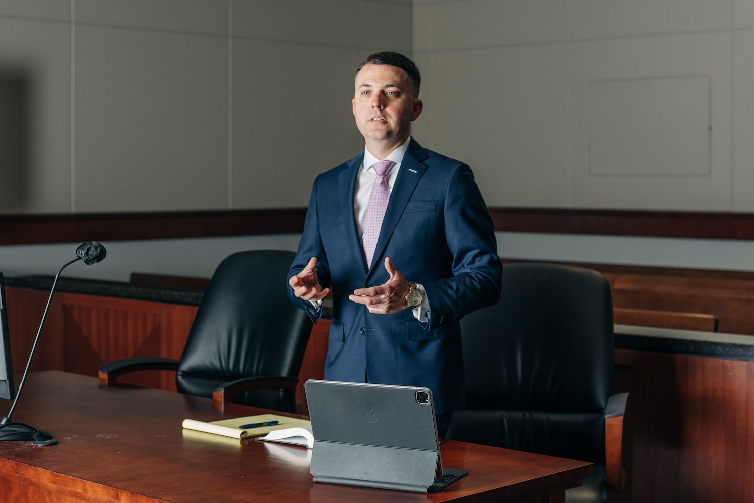 Man in a suit speaking at a courtroom podium with a tablet and notepad.