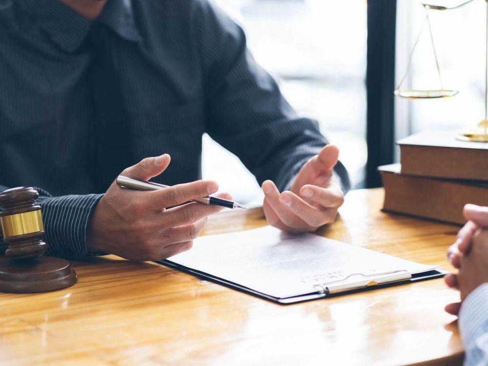 Legal consultation scene with a lawyer discussing documents at a desk, gavel and scales in view.