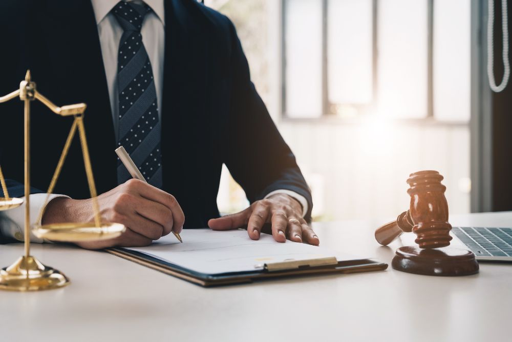 Lawyer signing documents at desk with gavel, scales, and laptop, symbolizing legal work and justice.