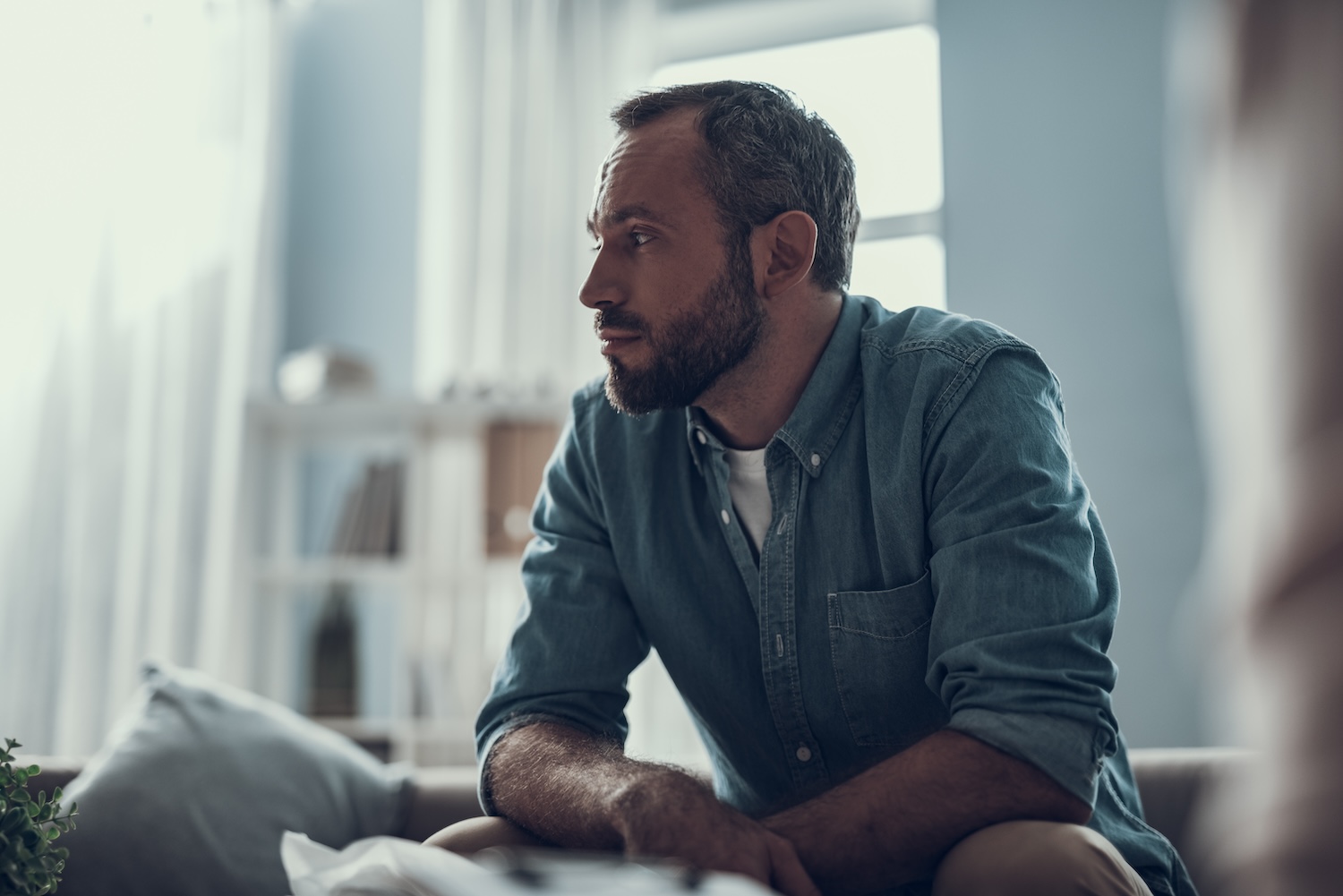 Man in denim shirt sitting thoughtfully in a well-lit room.