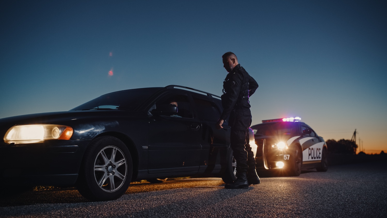 Police officer conducts traffic stop beside a car at dusk, with patrol lights illuminating the scene.